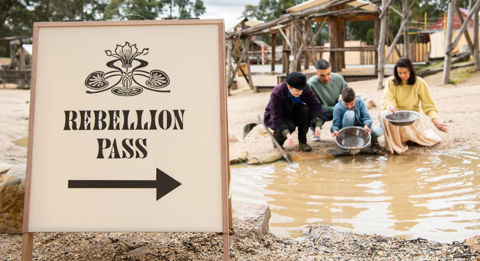 A sign reading "REBELLION PASS" with an arrow pointing right, and a family panning for gold in a shallow pool of water in a historical outdoor setting.