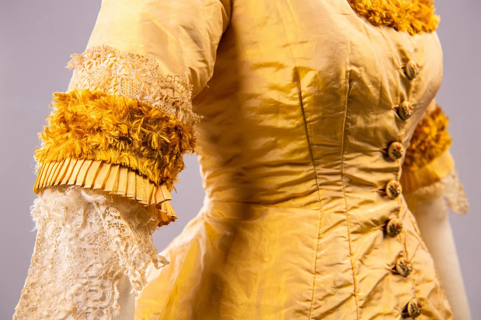 A close-up of a vintage yellow dress with ornate detailing, including lace sleeves, fringe trim, and decorative buttons, against a neutral background.