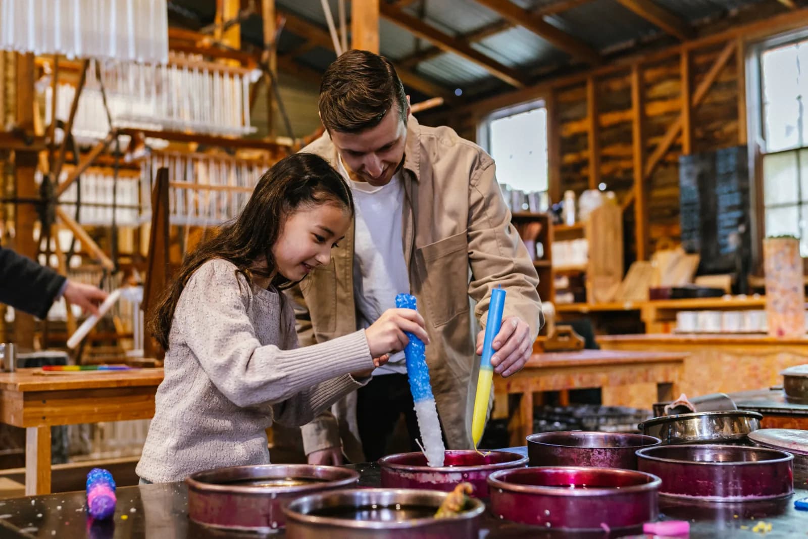 A young girl and a man dipping tools into bowls of colored substances in a workshop setting with wooden tables and equipment.