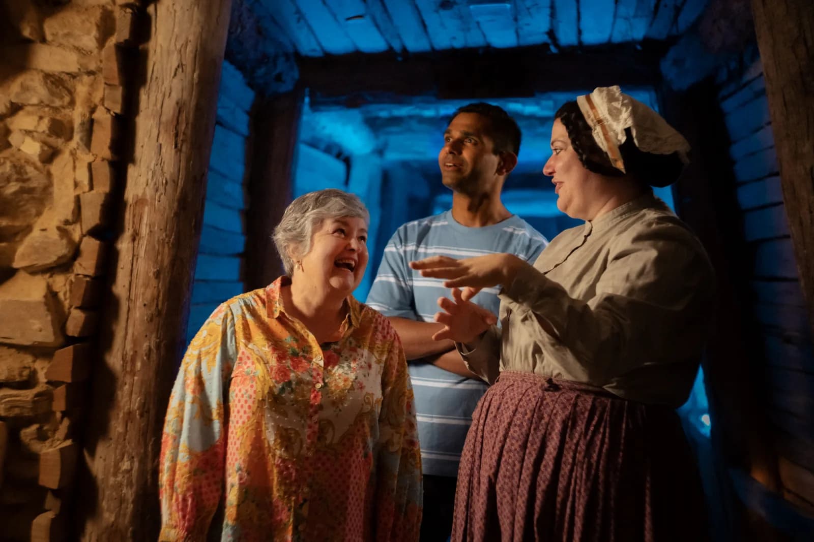 Three people stand inside a dimly lit, wooden tunnel with blue lighting. A woman in historical attire gestures while speaking to an older woman and a man in modern clothing, both smiling and engaged, suggesting a guided historical tour or interactive experience.