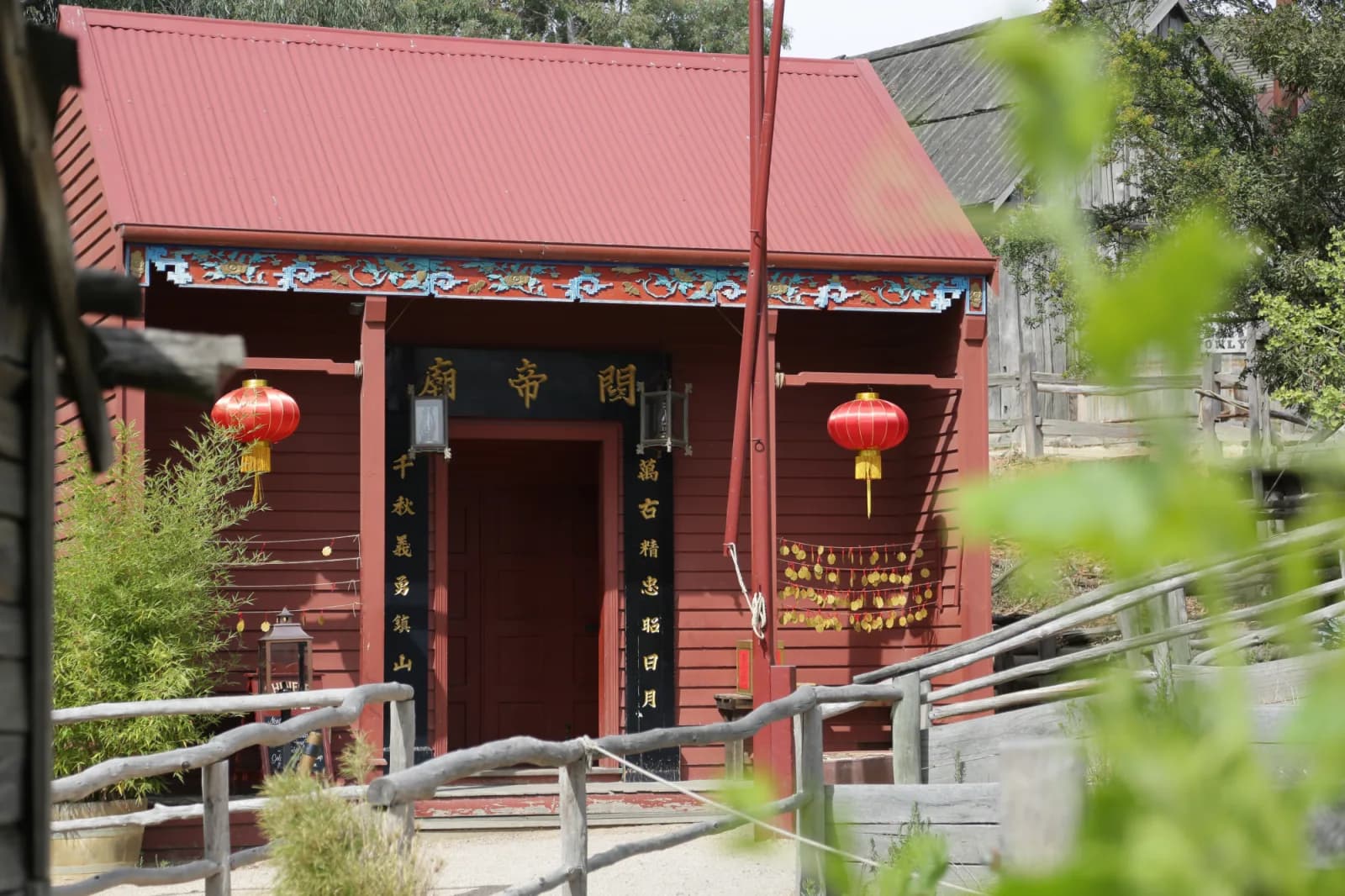 A small red building with a sloped roof features Chinese architectural elements, including hanging red lanterns and decorative trim with Chinese characters. The entrance is framed by wooden railings, and greenery is visible in the foreground, giving the scene a peaceful, cultural ambiance.