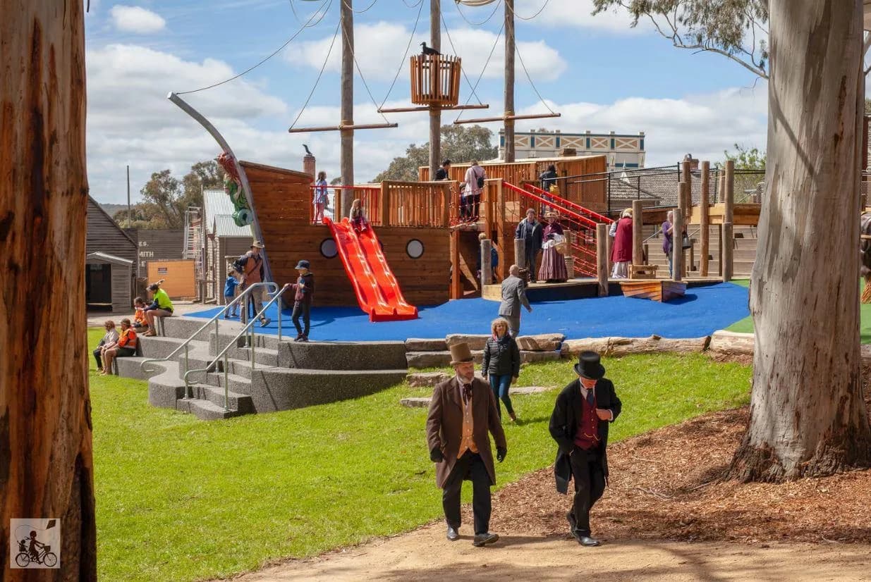 A wooden pirate ship playground with red slides is surrounded by children playing and adults supervising. The area has a blue safety surface, green grass, and tall trees under a partly cloudy sky. People walk along a dirt path in the foreground.