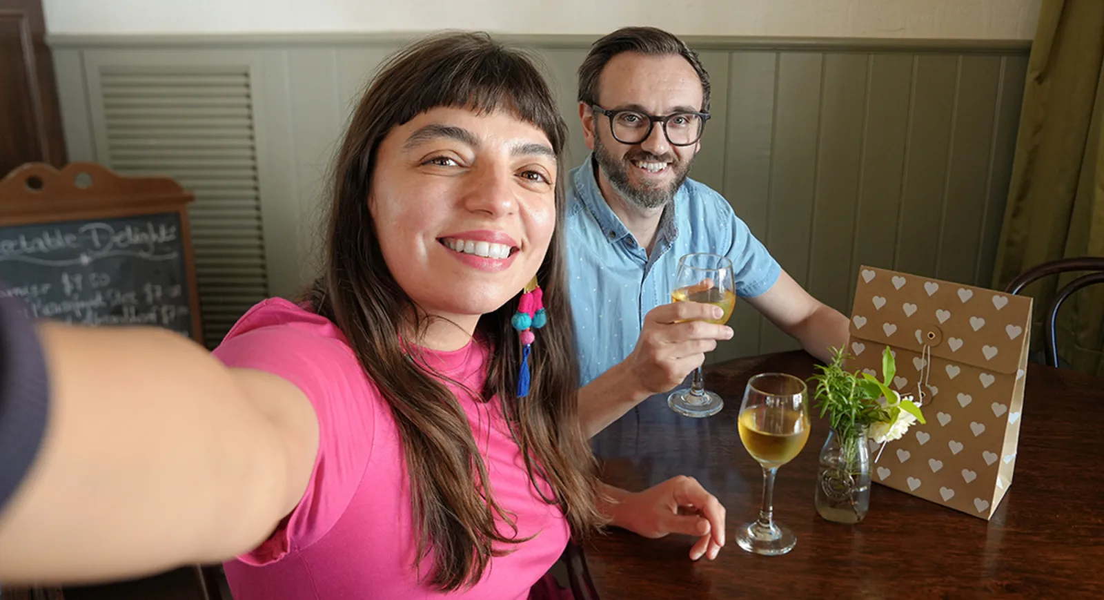 A cheerful couple enjoys a relaxed moment at a rustic table inside a historic building at Sovereign Hill, raising glasses of wine, with a gift bag and fresh herbs nearby, embodying the warm, immersive hospitality and heritage charm of Sovereign Hill.