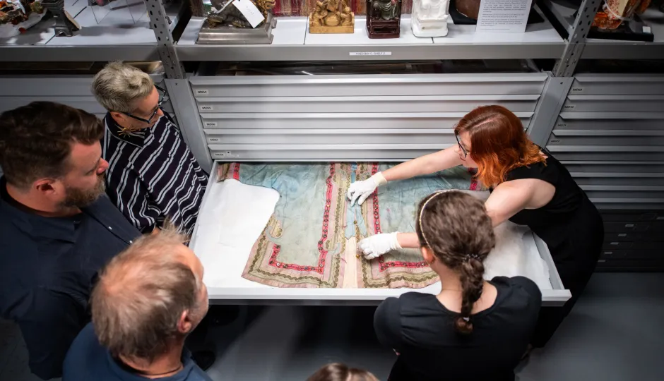 Museum conservators and visitors examining a historical garment with intricate embroidery on a pull-out storage shelf in Sovereign Hill