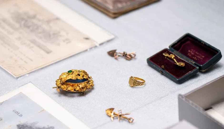 A gold brooch with a dark blue or green stone, a gold ring, two small gold pins or brooches, and a small jewelry box with a pin inside laid out on a grey surface