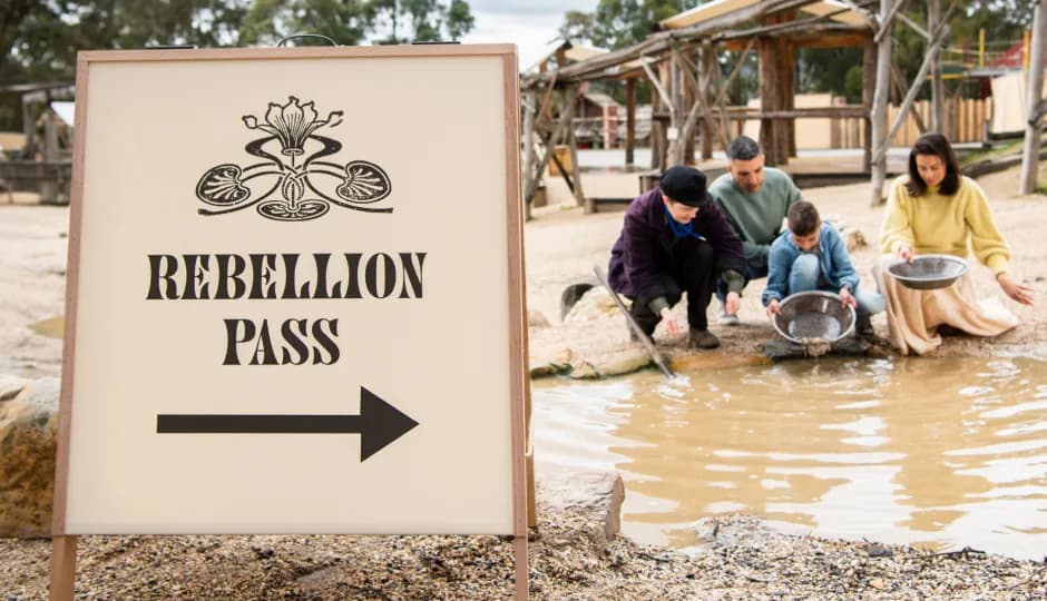 A sign reading "REBELLION PASS" with an arrow pointing right, and a family panning for gold in a shallow pool of water in a historical outdoor setting.