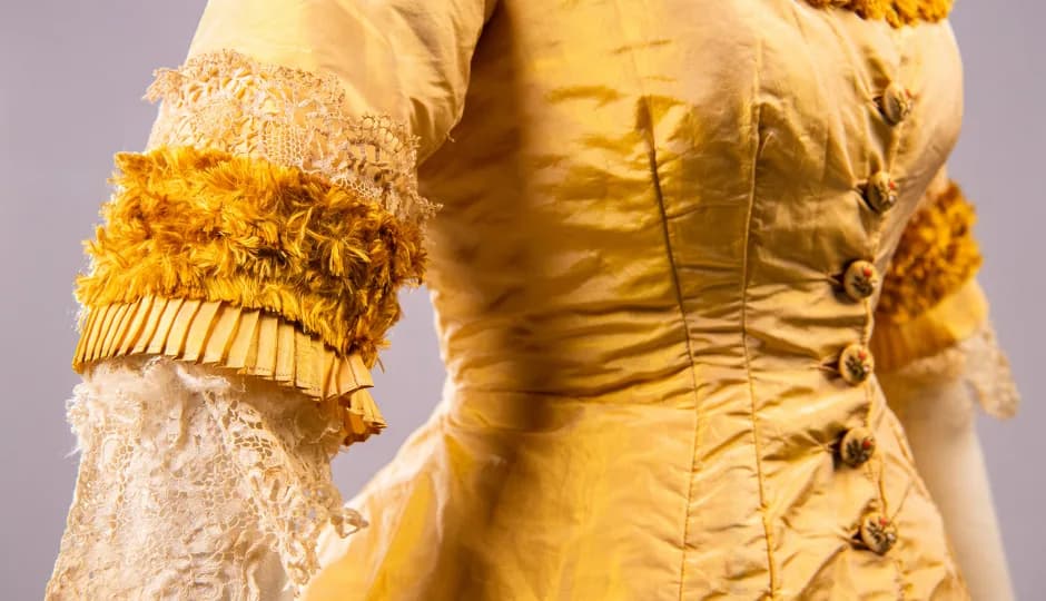 A close-up of a vintage yellow dress with ornate detailing, including lace sleeves, fringe trim, and decorative buttons, against a neutral background.