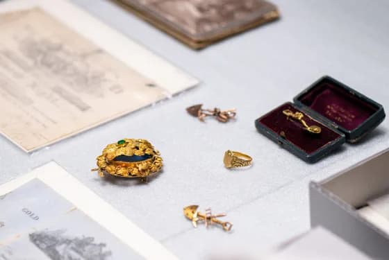 A gold brooch with a dark blue or green stone, a gold ring, two small gold pins or brooches, and a small jewelry box with a pin inside laid out on a grey surface