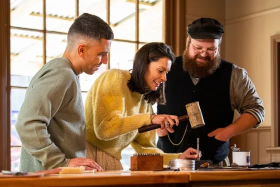 Three people are gathered around a wooden table, engaged in a hands-on activity. The person in the center, a woman with long dark hair, is using a mallet to work on a small object.