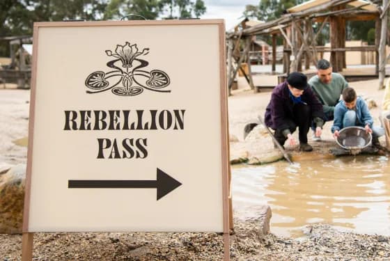 A sign reading "REBELLION PASS" with an arrow pointing right, and a family panning for gold in a shallow pool of water in a historical outdoor setting.