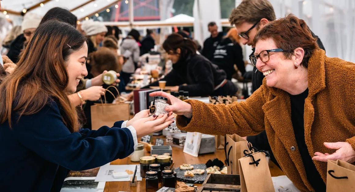 Two women at an indoor market stall, smiling and exchanging a small jar product, with other products and paper bags on the table.