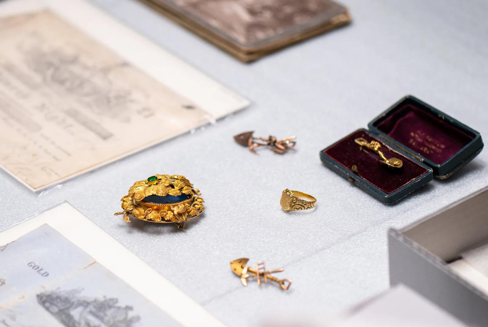 A gold brooch with a dark blue or green stone, a gold ring, two small gold pins or brooches, and a small jewelry box with a pin inside laid out on a grey surface