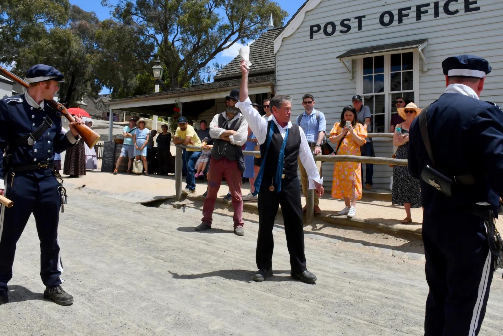 A scene depicting a historical reenactment or performance in front of a building labeled "POST OFFICE." A man in the center is gesturing with a raised hand, seemingly addressing a crowd of people. Two men dressed in dark blue uniforms, one holding a rifle, stand at the edges of the scene. The setting suggests a historical village or outdoor museum.