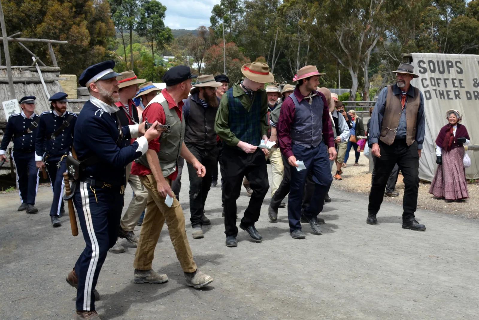 A group of people dressed in historical clothing are walking along a dirt road, with a sign in the background advertising "Soups, Coffee & Other Drinks." The people appear to be participating in a historical reenactment or are part of a themed event. The setting suggests an outdoor historical site or village.