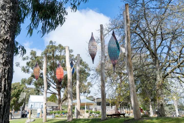 Outdoor art installation with colorful hanging lanterns suspended from tall wooden posts among trees and a building in the background.