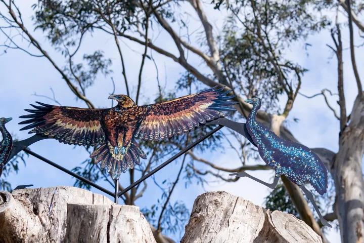 A metal sculpture of an eagle with outstretched wings and patterned feathers, perched on a wooden post with tree branches and a blue bird sculpture in the background.