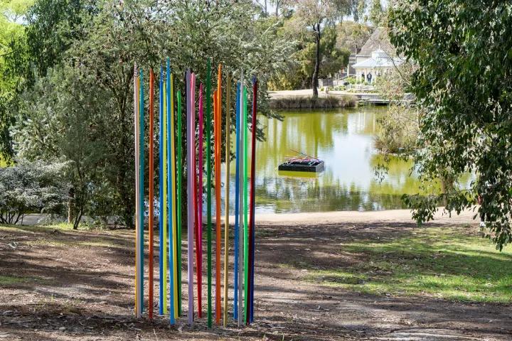 A group of tall, thin, colorful sticks arranged vertically in a natural outdoor setting with trees and foliage in the background.