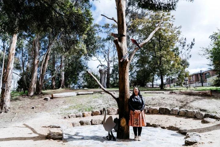 A woman in a dark jacket and patterned skirt standing next to a tree with bird sculptures attached to its branches, in an outdoor setting with a circular rock border and trees in the background.
