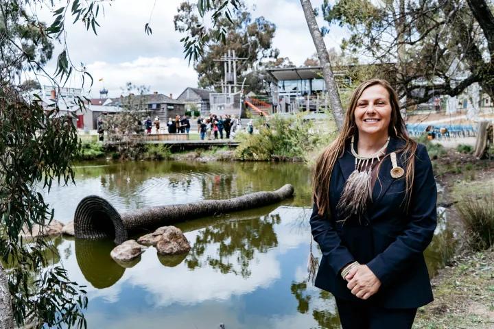 A woman with long brown hair, wearing a dark blazer and a necklace with feathers, standing in front of a pond with a submerged sculpture and buildings in the background.