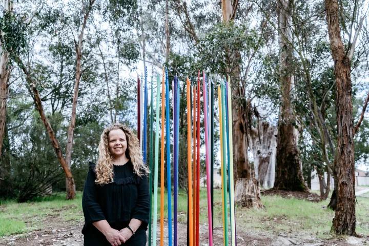 A woman with curly blonde hair, wearing a black dress, standing in front of a colorful vertical stick installation in a wooded area with tall trees.