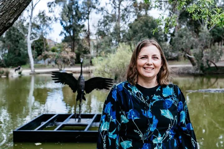 A woman with shoulder-length brown hair and a blue patterned top smiles at the camera, with a pond, trees, and a black metal crane sculpture in the background.