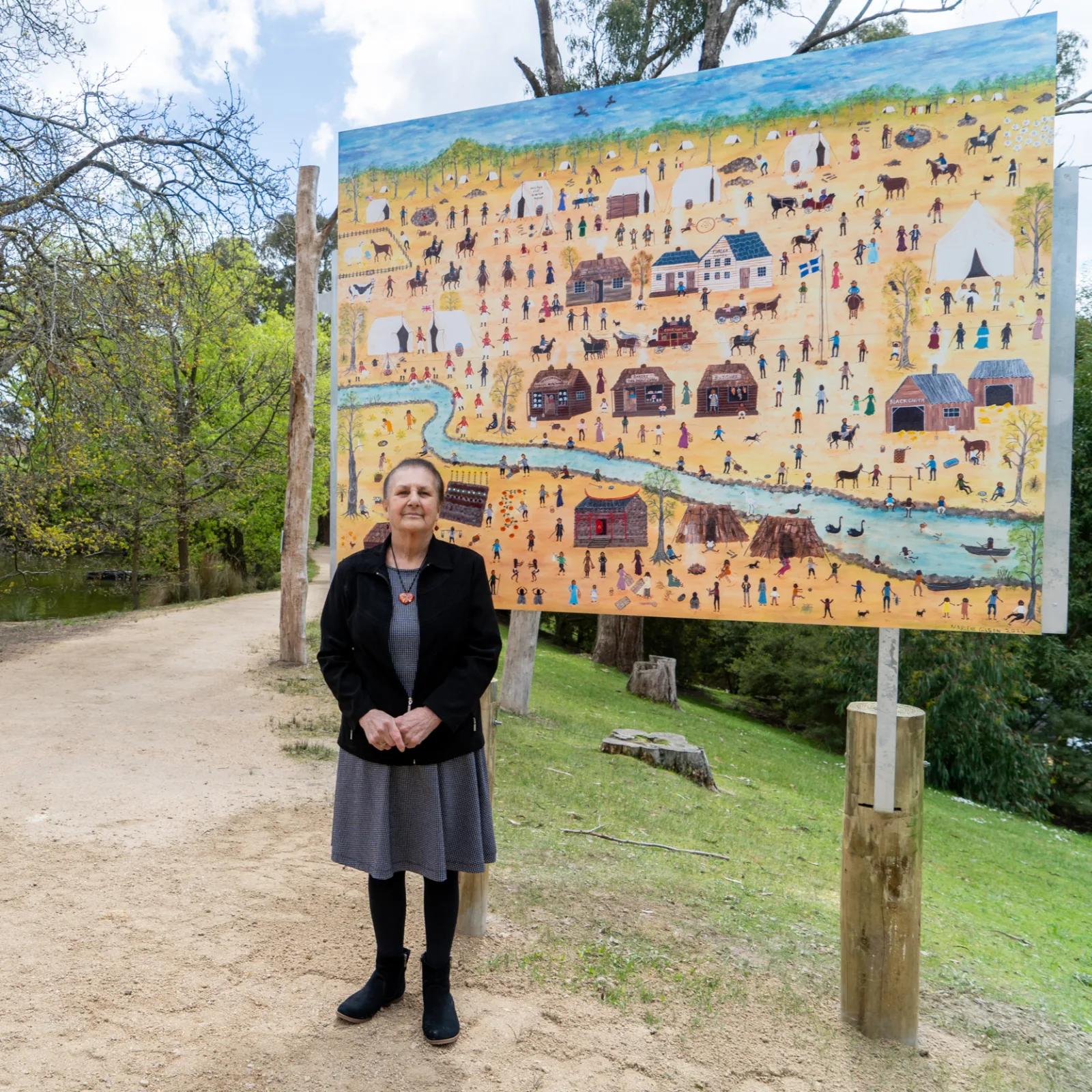 Marlene Gilson standing next to a large painted canvas depicting a village or landscape, displayed outdoors on a stand in a park setting.