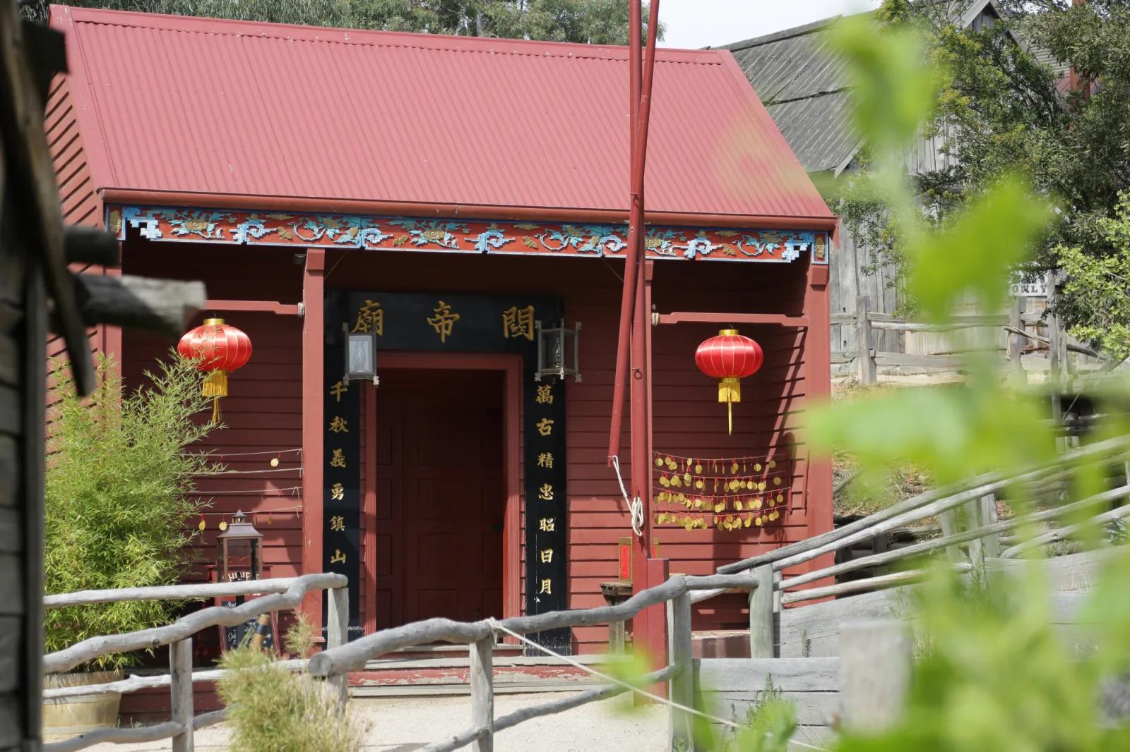 A small red building with a sloped roof features Chinese architectural elements, including hanging red lanterns and decorative trim with Chinese characters. The entrance is framed by wooden railings, and greenery is visible in the foreground, giving the scene a peaceful, cultural ambiance.