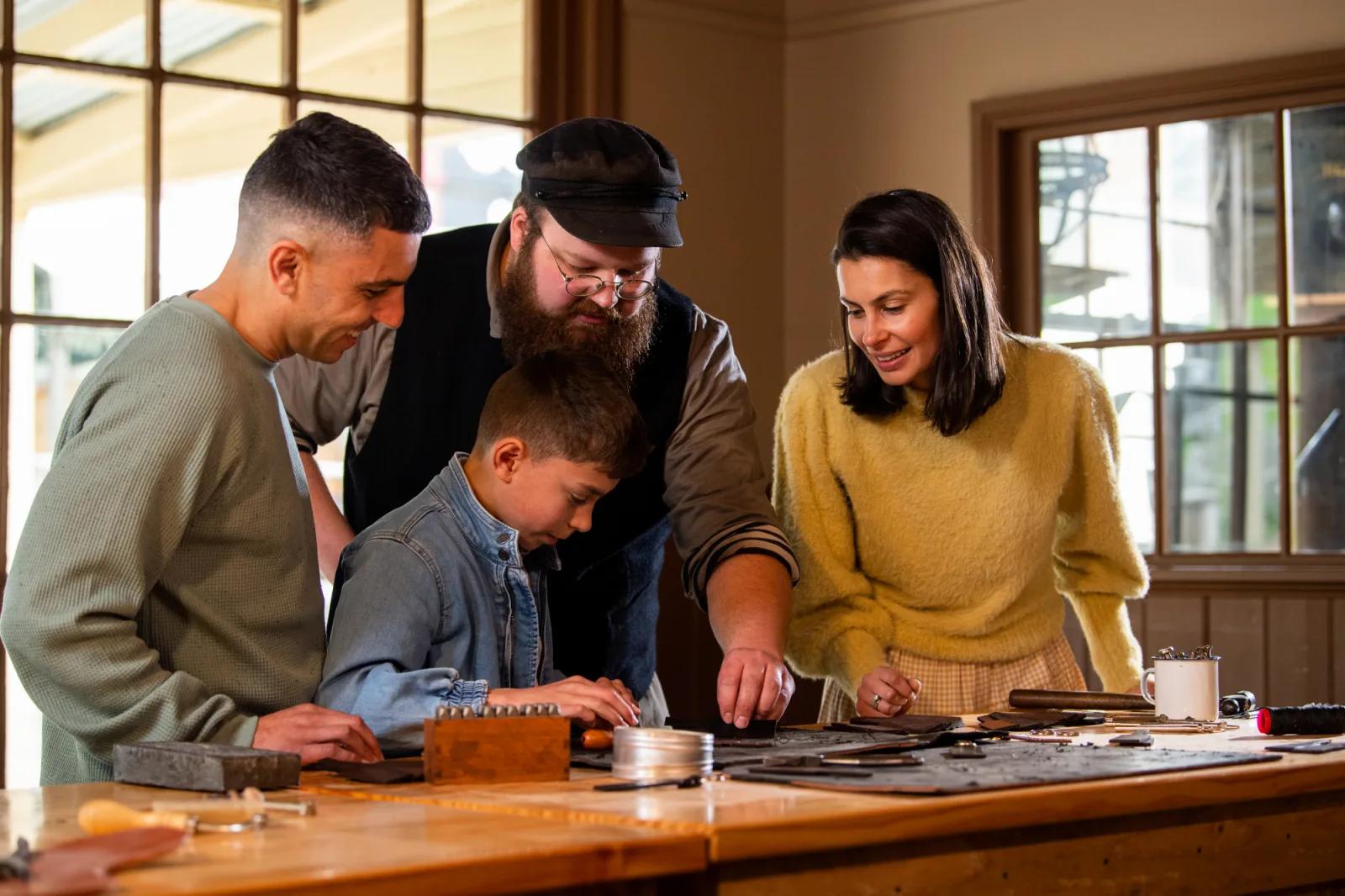 A family of three, along with an instructor in vintage attire, are gathered around a wooden workbench inside a well-lit room. The instructor is demonstrating a craft, possibly metal or leatherworking, while the child focuses on the project. Various tools and materials are spread across the table.