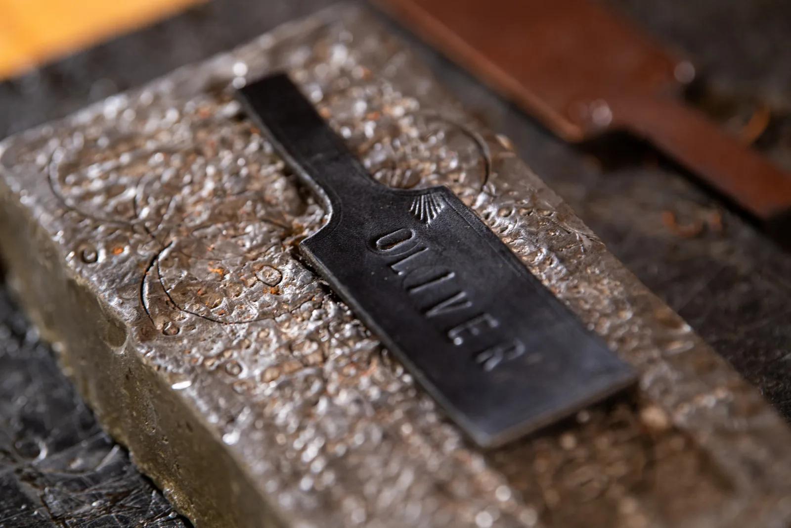A close-up of a metal tag with the name OLIVER stamped on it, resting on a textured metal block. The tag has a rectangular shape with a small handle-like extension at one end, likely part of a metalworking or engraving project.