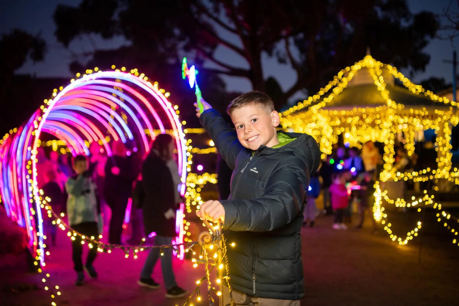 Young boy posing with glowing wand inside illuminated tunnel and festive light displays, from Australia’s most visited outdoor museum and heritage attraction.