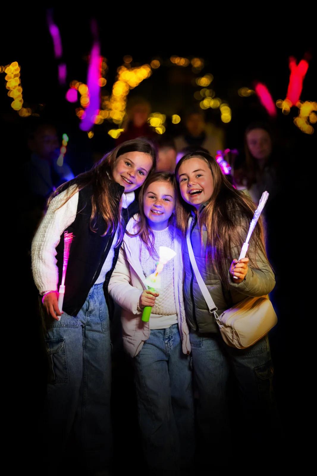 Three smiling girls holding glowing light wands during nighttime festivities, from Australia’s most visited outdoor museum and heritage attraction.