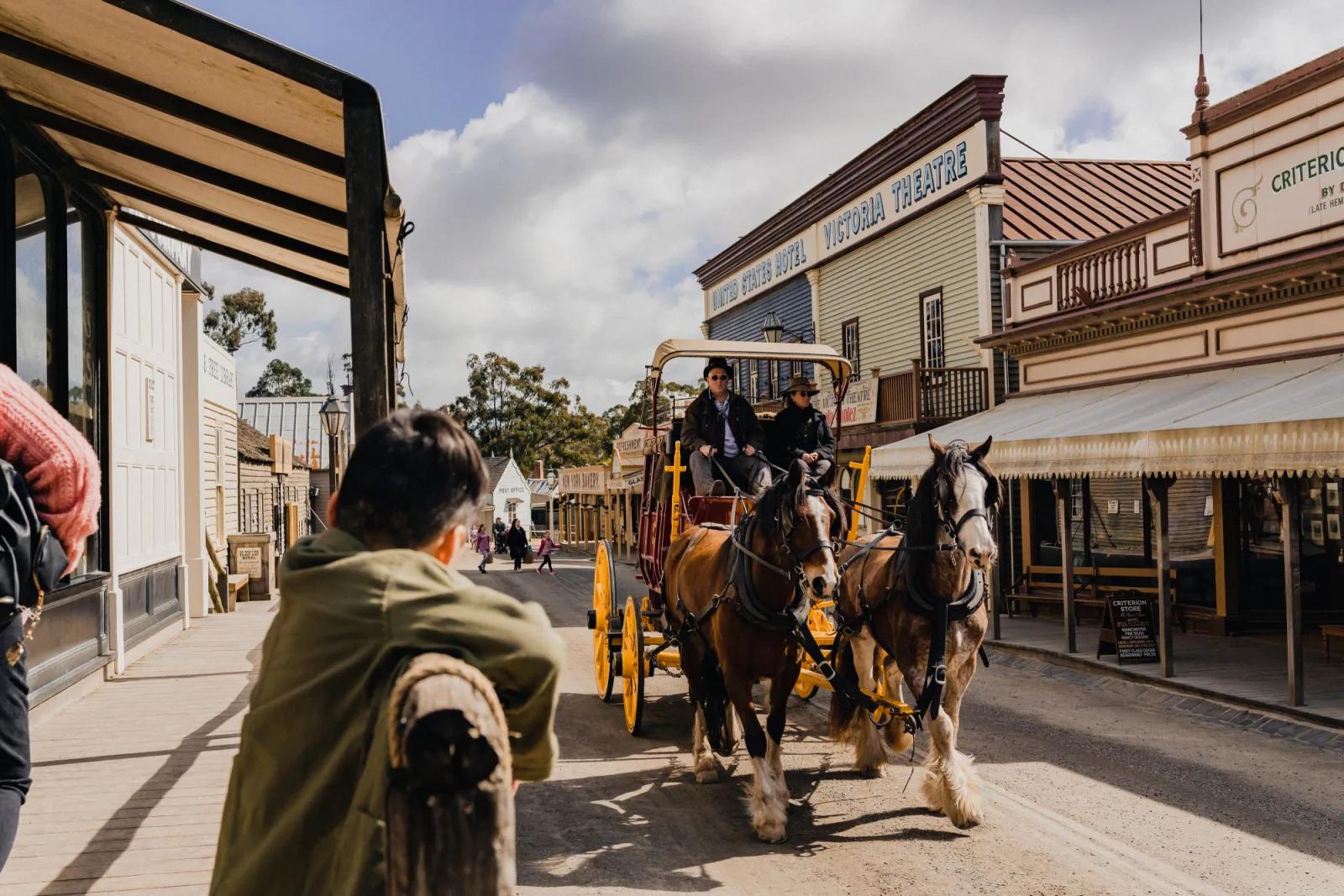 A horse-drawn carriage with two passengers rolls down the historic main street of Sovereign Hill, passing iconic buildings including the United States Hotel, Victoria Theatre, and Criterion Store. Visitors watch from the wooden verandahs under a partly cloudy sky capturing the immersive, living-history atmosphere of Sovereign Hill.