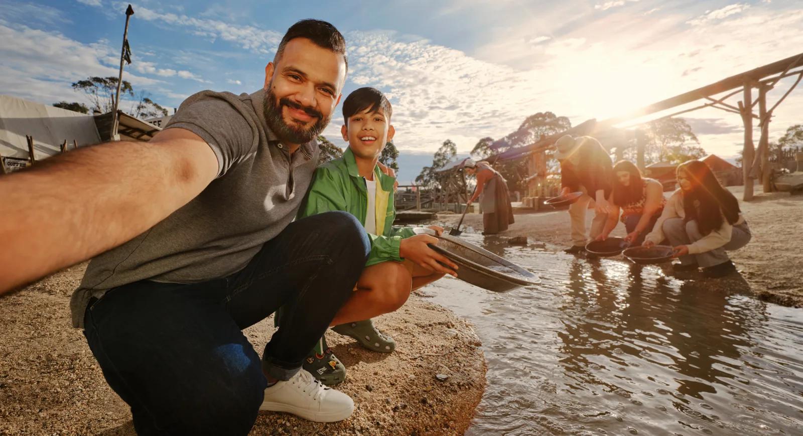 At Sovereign Hill, a man and boy capture a joyful selfie while gold panning along a sunlit creek, surrounded by other visitors engaged in the hands-on activity. Historic gold rush-era buildings line the background under a bright blue sky, highlighting the interactive, family-friendly charm of Sovereign Hill’s living history experience.