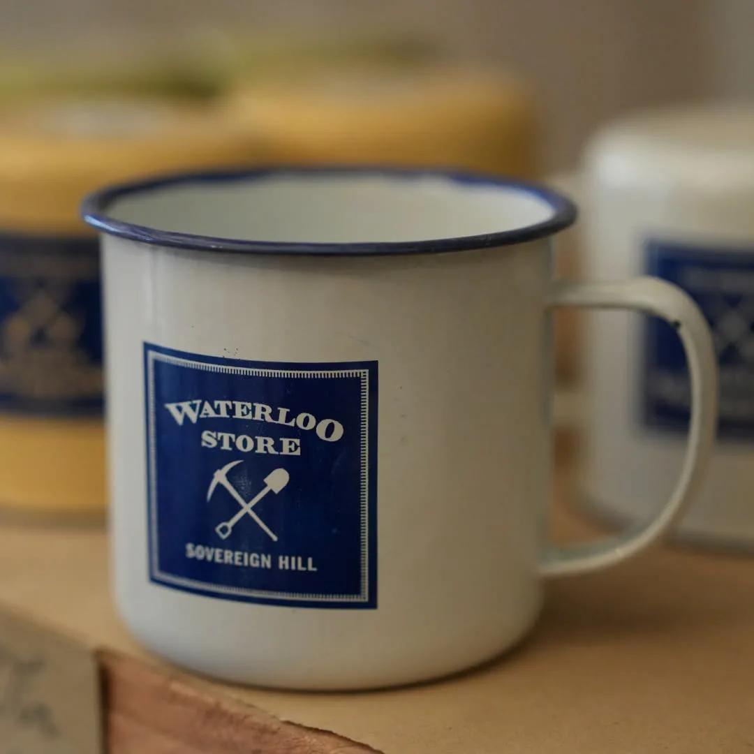 Cream enamel mug featuring the Waterloo Store and Sovereign Hill logo, displayed among matching merchandise.