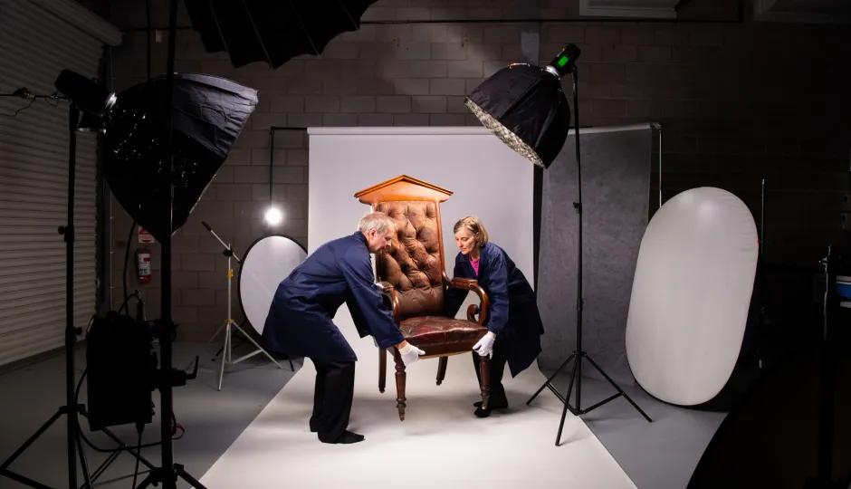 Two people in blue protective smocks and white gloves carefully moving a vintage upholstered armchair on a white backdrop, surrounded by professional photography studio lighting and equipment in Sovereign Hill