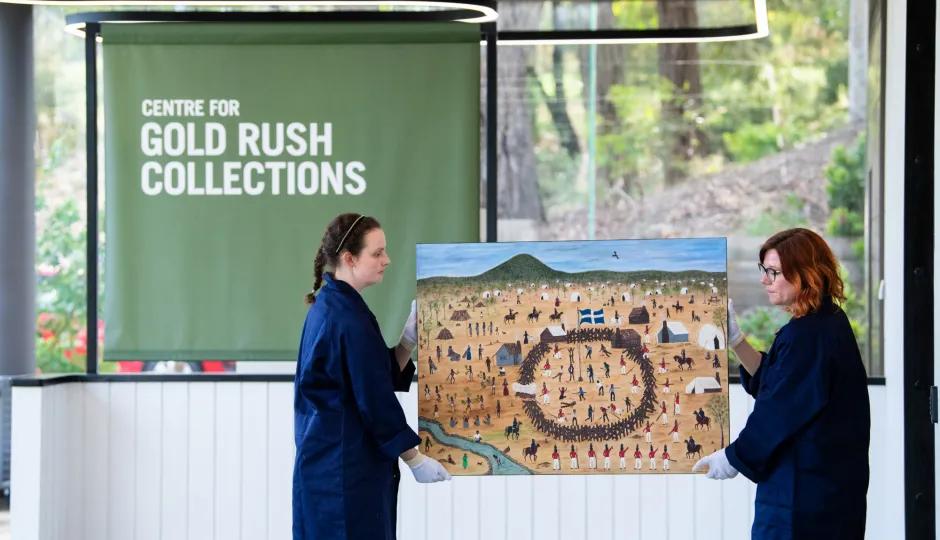 Two people in dark blue smocks and white gloves carefully holding a large, colorful painting depicting a historical scene with numerous figures, horses, and flags, in front of a green banner that reads 'CENTRE FOR GOLD RUSH COLLECTIONS'.