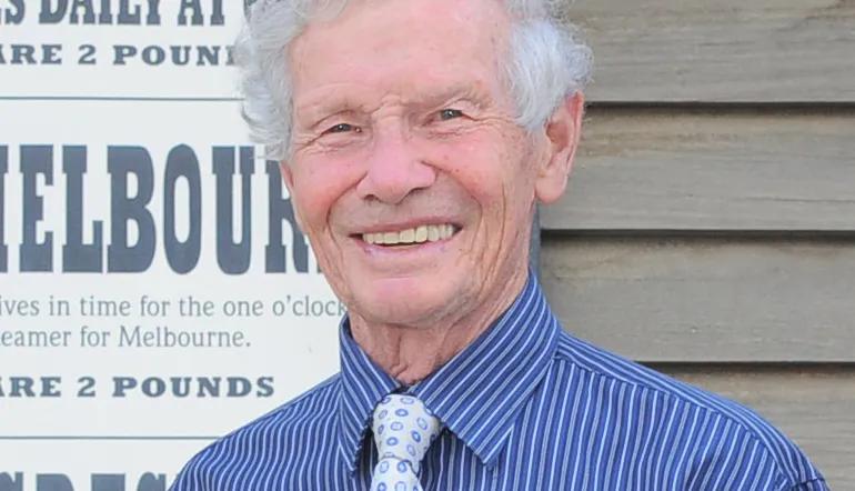 Close-up of a smiling older man with white hair, wearing a striped blue shirt and a patterned tie.