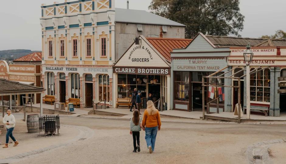 Visitors stroll down the authentic, dust-dusted Main Street at Sovereign Hill, passing historic storefronts including Clarke Brothers Wholesale & Family Grocers, Ballarat Times – C. Spencer, T. Murphy, California Tent Maker, and Rees & Benjamin Watch & Clock Makers. A mother and daughter walk hand-in-hand toward the shops, embodying the immersive, family-friendly experience of Sovereign Hill’s living gold rush town often referred to as Main Street min in promotional contexts for its miniature-scale authenticity and charm.