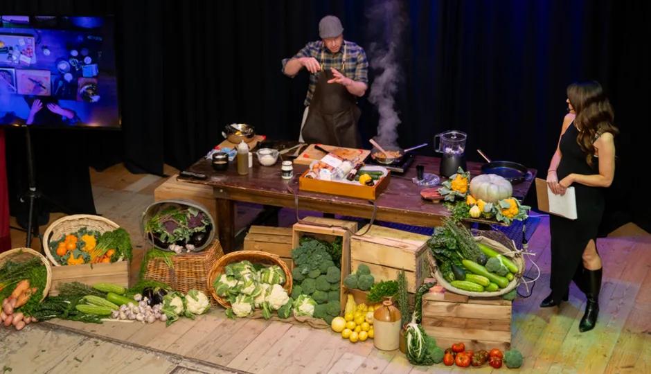 At Sovereign Hill, a dynamic live cooking demonstration unfolds on stage, where a chef in a plaid shirt and apron prepares a dish amid an abundant display of fresh produce, including carrots, broccoli, pumpkins, and leafy greens, arranged in wooden crates and woven baskets. A host in a black dress observes with a clipboard, while a screen projects the action, capturing the interactive and immersive culinary experience that brings heritage-inspired cooking to life at Sovereign Hill.