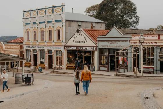 Visitors stroll down the authentic, dust-dusted Main Street at Sovereign Hill, passing historic storefronts including Clarke Brothers Wholesale & Family Grocers, Ballarat Times – C. Spencer, T. Murphy, California Tent Maker, and Rees & Benjamin Watch & Clock Makers. A mother and daughter walk hand-in-hand toward the shops, embodying the immersive, family-friendly experience of Sovereign Hill’s living gold rush town often referred to as Main Street min in promotional contexts for its miniature-scale authenticity and charm.