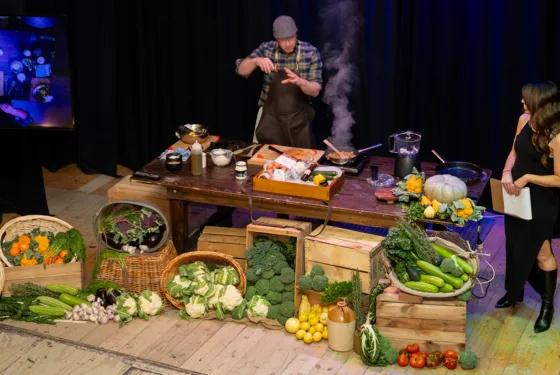 At Sovereign Hill, a dynamic live cooking demonstration unfolds on stage, where a chef in a plaid shirt and apron prepares a dish amid an abundant display of fresh produce, including carrots, broccoli, pumpkins, and leafy greens, arranged in wooden crates and woven baskets. A host in a black dress observes with a clipboard, while a screen projects the action, capturing the interactive and immersive culinary experience that brings heritage-inspired cooking to life at Sovereign Hill.