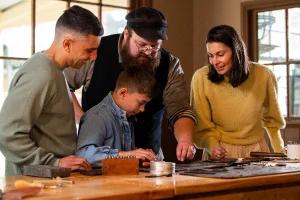 A group of four people, including a man with a beard and a cap, a boy, a man with a shaved head, and a woman in a yellow sweater, working together at a table with tools and materials.