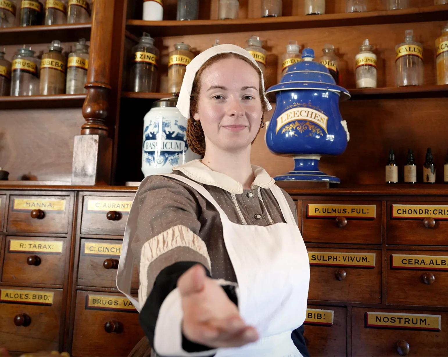 Costumed apothecary reenactor extending a welcoming hand inside a historic pharmacy, from Australia’s most visited outdoor museum and heritage attraction.