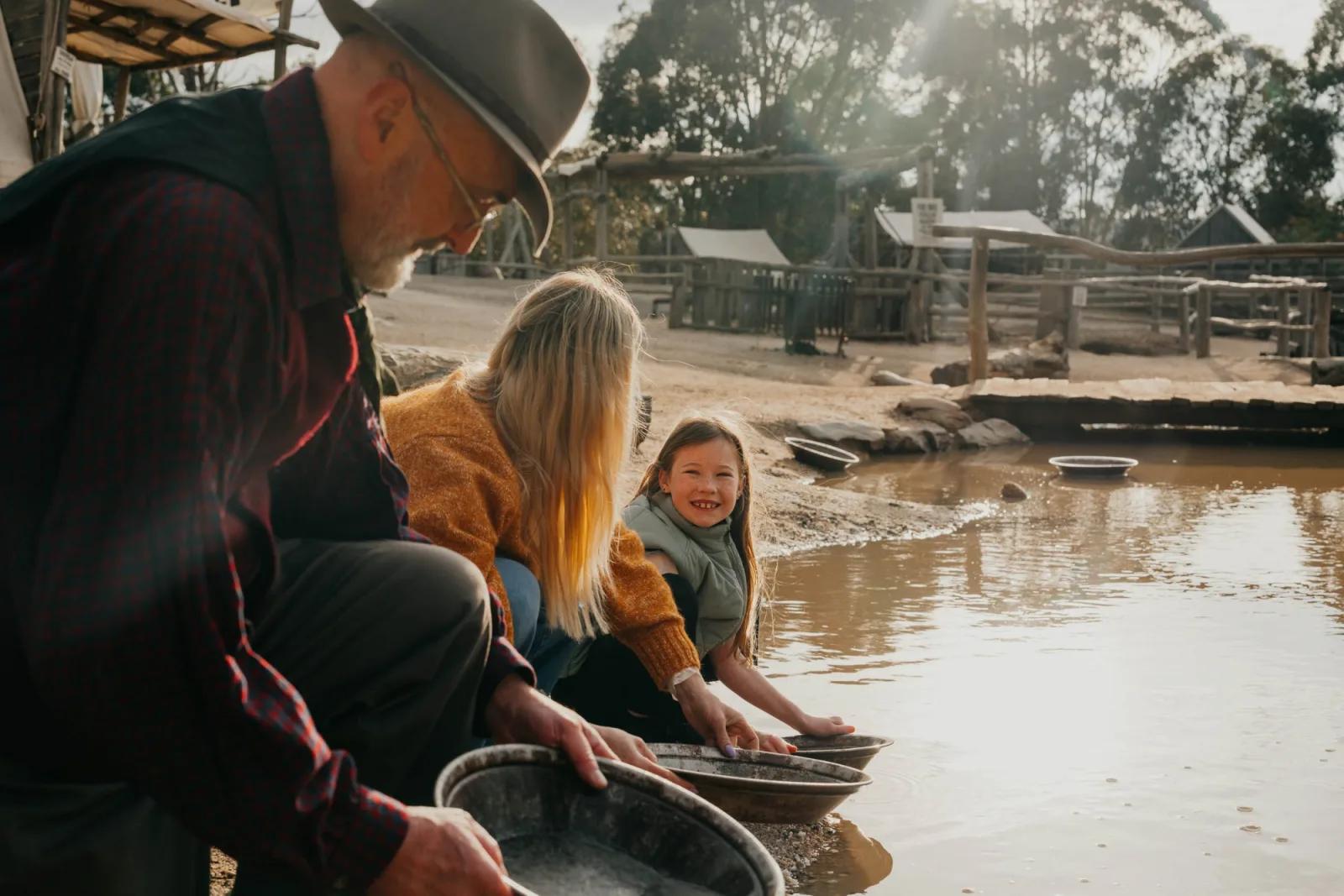 A family enjoys Gold Panning at Sovereign Hill, guided by a costumed interpreter in period attire as they swirl water and sediment in metal pans beside a sun-dappled creek. The young girl smiles brightly, fully immersed in the hands-on experience, while rustic tents and wooden structures of the historic gold rush town stand in the background capturing the joy and authenticity of Sovereign Hill’s signature activity.