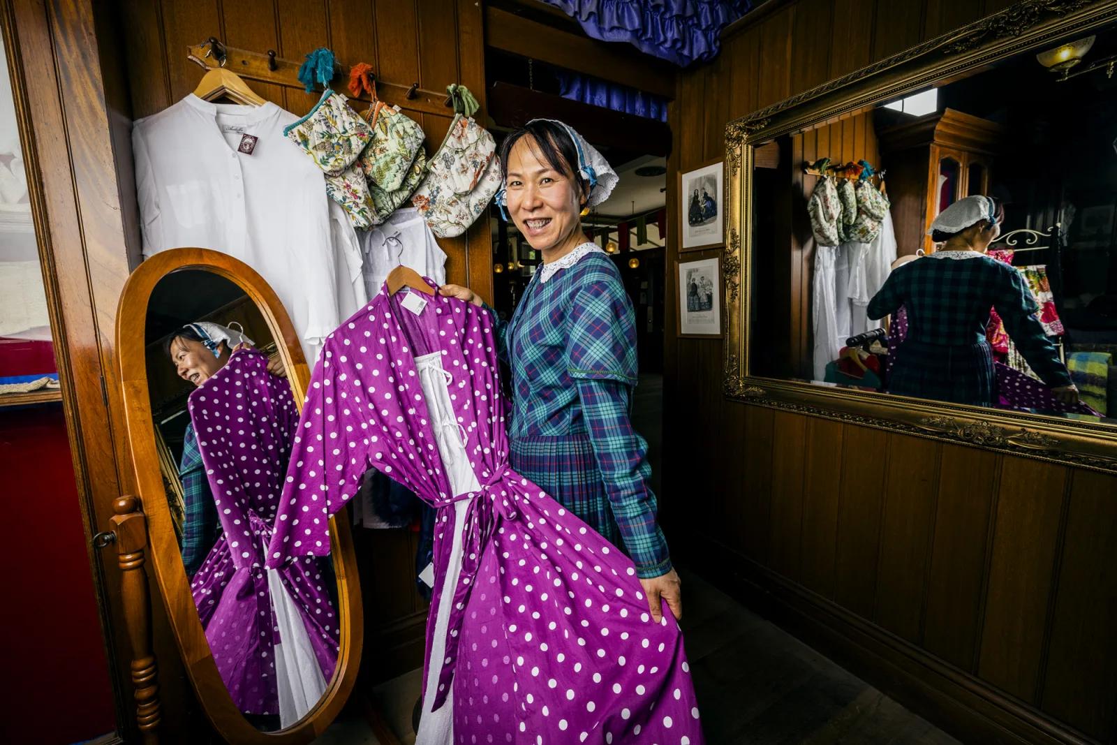 A smiling visitor tries on a vibrant purple polka-dot dress inside a charming 19th-century-style shop at Sovereign Hill, surrounded by vintage garments and wooden mirrors, capturing the fun, interactive fashion experience that brings gold rush history to life at Sovereign Hill.