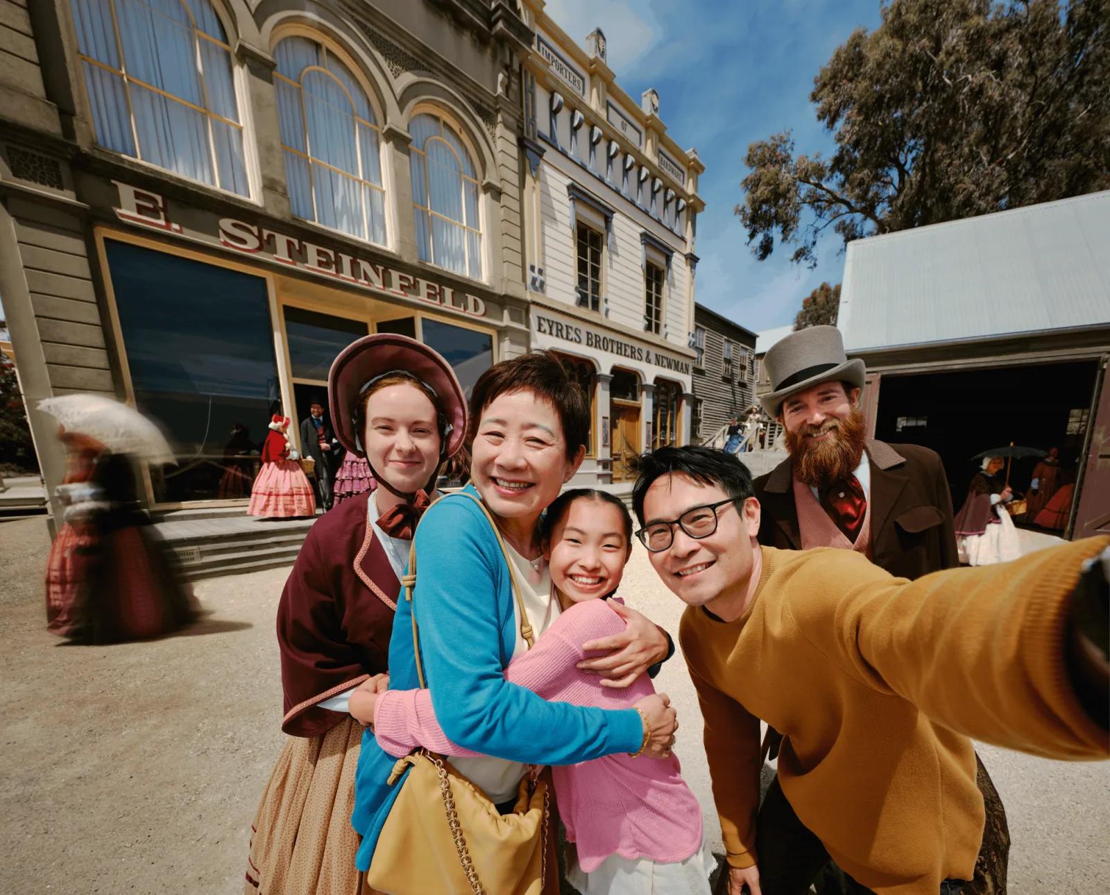 A joyful group captures a selfie in front of historic gold rush-era buildings at Sovereign Hill, including E. Steinfeld and EYRES BROTHERS & NEWMAN, as costumed interpreters in period attire, including a woman in a pink dress and a bearded man in a top hat, enhance the immersive atmosphere. Sunlight bathes the vibrant street, highlighting Sovereign Hill’s authentic 1850s charm and the seamless blend of visitor experience with living history.