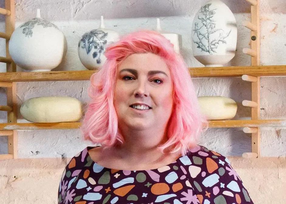 Portrait of a woman with pink hair standing in front of ceramic vessels displayed on wooden shelves at Sovereign Hill.