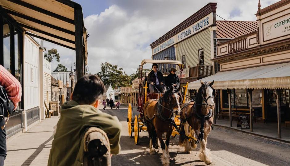A horse-drawn carriage with two passengers rolls down the historic main street of Sovereign Hill, passing iconic buildings including the United States Hotel, Victoria Theatre, and Criterion Store. Visitors watch from the wooden verandahs under a partly cloudy sky capturing the immersive, living-history atmosphere of Sovereign Hill.