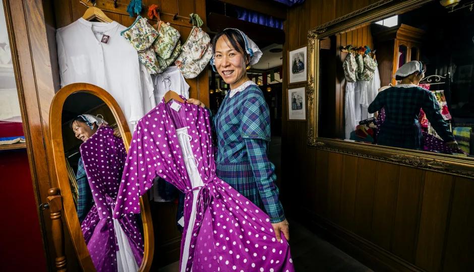 A smiling visitor tries on a vibrant purple polka-dot dress inside a charming 19th-century-style shop at Sovereign Hill, surrounded by vintage garments and wooden mirrors, capturing the fun, interactive fashion experience that brings gold rush history to life at Sovereign Hill.