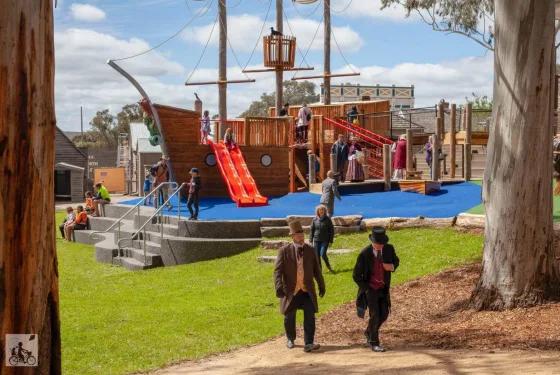A wooden pirate ship playground with red slides is surrounded by children playing and adults supervising. The area has a blue safety surface, green grass, and tall trees under a partly cloudy sky. People walk along a dirt path in the foreground.